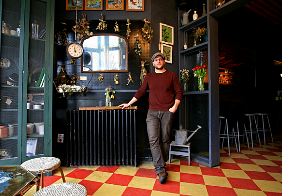 Justin Israelson at Sycamore bar, circa 2012, standing behind black counter with decorative hanging plants and mirror...